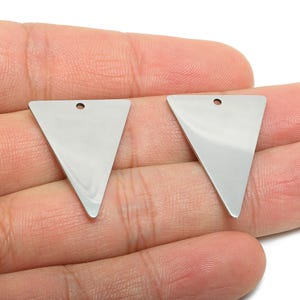 Puede incluir: Dos colgantes triangulares plateados y brillantes, con un pequeño agujero en la parte superior para ensartar. Los colgantes se sostienen en una mano, mostrando su superficie pulida y reflectante. Adecuado para la creación de joyas.