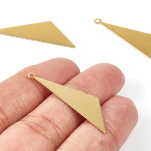 Peut inclure: Trois breloques géométriques dorées en forme de triangle avec une petite boucle en haut pour la fixation aux bijoux. Une breloque est tenue dans une main, tandis que les deux autres sont sur fond blanc.