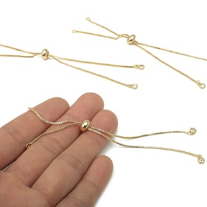 Pode incluir: Ajustadores de pulseira dourados com um design de contas deslizantes e corrente delicada. Os ajustadores têm pequenos laços nas extremidades para fixar à joia. Várias peças sobre fundo branco.