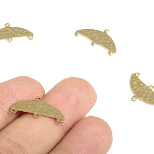 Puede incluir: Varios hallazgos de joyería dorados en forma de media luna con una textura martillada. Cada hallazgo tiene tres pequeños bucles para sujetar a otros componentes. Un hallazgo se sostiene en una mano, otros están dispersos sobre una superficie blanca.