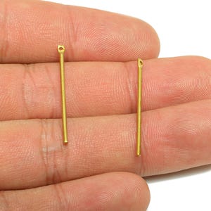 May include: Two gold-colored, straight head pins with a loop at the top. The pins are held in a hand against a white background. These jewelry-making components are used for crafting earrings, necklaces, and other accessories.