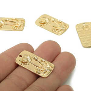 Puede incluir: Encantos rectangulares de metal dorado con un diseño de flor de amapola. Los encantos tienen un patrón floral en relieve detallado, que incluye flores y tallos de amapola. Un encanto se sostiene en una mano.