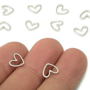 May include: Silver-colored heart-shaped charms. The charms are small and delicate, with a simple outline design. Several charms are scattered on a white background, with a few held between fingers. These charms are suitable for jewelry making.