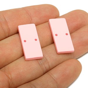 以下が含まれることがあります： 2つの薄いピンク色の長方形のドミノで、それぞれに2つの小さな赤い点があります。ドミノは手で持たれており、背景は白です。ドミノは単色で、プラスチックまたは樹脂でできているようです。