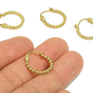 Puede incluir: Pendientes de aro dorados con diseño de cuentas. Un pendiente se sostiene en una mano, con tres pendientes adicionales visibles. Los pendientes tienen un diseño simple y elegante, perfecto para el uso diario. Los pendientes son pequeños y ligeros.