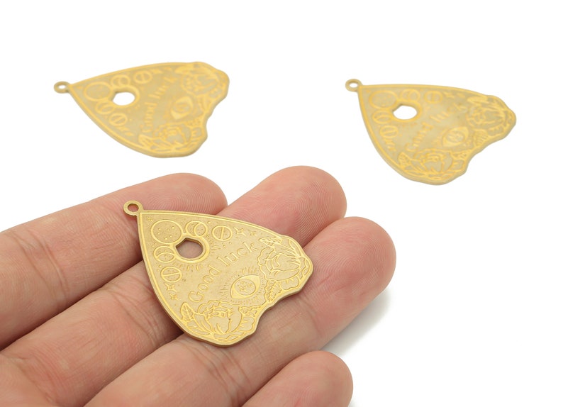May include: Three gold-colored metal charms shaped like a triangle with a hole in the center. The charms have an etched design of a circle with a star inside, a crescent moon, and the words "Good Luck".