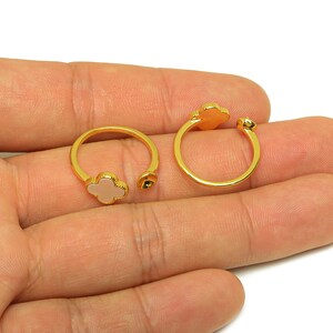 May include: Two gold-colored open rings with a clover-shaped design and a light pink center. The rings are open-ended, allowing for size adjustment. The rings are on a white background.