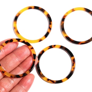 May include: Four round, open-center rings with a tortoiseshell pattern of brown and yellow-orange. One ring is held in a hand, while the other three are scattered on a white surface. The rings are likely jewelry components or craft supplies.