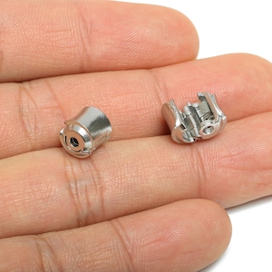 May include: Two small, shiny, silver-coloured metal objects. One is a cylindrical shape with a hole in the centre, and the other is a more complex, open design. The objects are held in a hand, suggesting a small size.