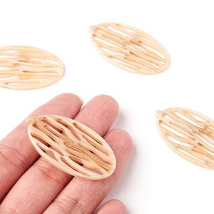 Peut inclure: Quatre pendentifs ovales beiges avec un motif tissé. L'un des pendentifs est tenu dans une main. Les pendentifs ont une petite boucle en haut pour l'enfilage. Le fond est blanc.