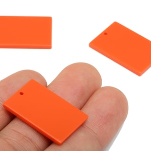 Puede incluir: Tres colgantes rectangulares de color naranja brillante con un pequeño agujero en la parte superior para ensartar. Un colgante se sostiene en una mano, mientras que otros dos se muestran por separado sobre un fondo blanco. Estos colgantes son ideales para la fabricación de joyas.