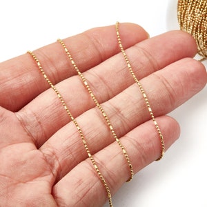 May include: A close-up of a gold chain with small, round beads. The chain is lying on a hand, and the background is white.