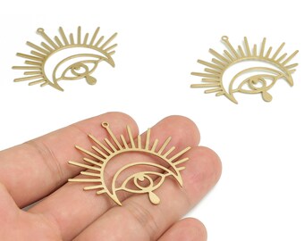 May include: Gold-colored metal eye charm with sunburst design. The charm features an eye with a teardrop and a crescent moon shape. The charm has a small loop at the top for attaching to jewelry. The image shows three charms.