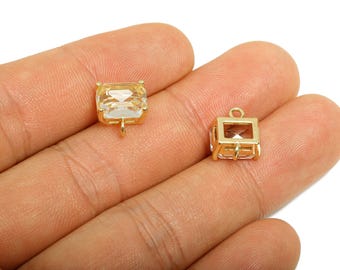 Puede incluir: Dos colgantes cúbicos dorados con cristales rectangulares transparentes. Un colgante tiene un cristal sólido, el otro un marco hueco. Ambos tienen pequeños bucles para sujetarlos a la joyería. Los colgantes se sostienen en una mano.