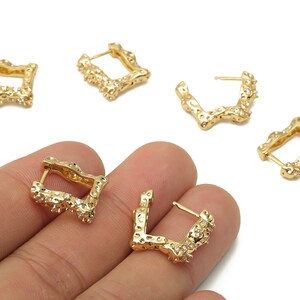May include: Gold-toned square hoop earrings with a textured, organic design. The earrings feature small, clear crystals and a post closure. Several earrings are displayed on a white background, with some held in a hand.
