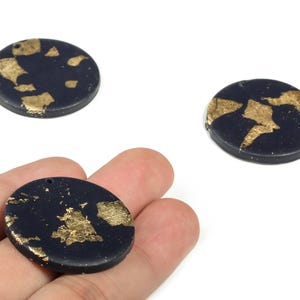 May include: Three dark blue, round polymer clay beads with gold leaf accents. One bead is held in a hand. Each bead has a small hole for stringing. The beads are on a white background.