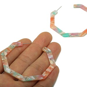 May include: A pair of geometric hoop earrings. The earrings are a marbled pattern of pink, orange, white, and teal. The earrings are held in a hand, with one earring in the background.