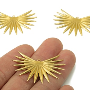 May include: Three gold-colored metal pendants in a sunburst design. One pendant is held in a hand, while the other two are displayed above. Each pendant has a small hole at the top for stringing.