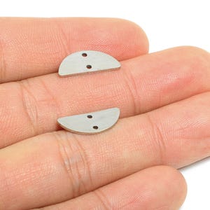 May include: Two silver-coloured, half-moon shaped metal jewellery findings with two holes each. The findings are displayed on a hand, and are likely used for jewellery making or crafting. The metal has a brushed finish.
