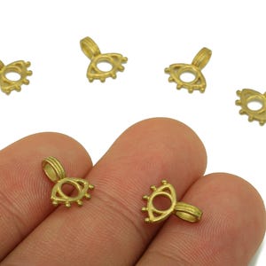 May include: Several small, gold-colored eye-shaped jewelry findings. The findings have a loop at the top for attaching to a chain or other jewelry component. The eye shape has a central hole and small decorative details.