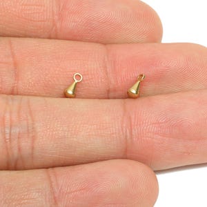 Könnte beinhalten: Zwei kleine, goldfarbene Tropfenanhänger. Jeder Anhänger hat oben eine Schlaufe zum Befestigen an Schmuck. Die Anhänger werden vor einem hautfarbenen Hintergrund gezeigt.