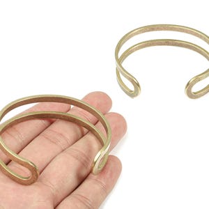 Puede incluir: Dos pulseras de puño doradas. Una pulsera se sostiene en una mano, mientras que la otra se muestra por separado. Cada pulsera presenta un diseño de doble banda con extremos curvos y abiertos. Las pulseras son de metal.