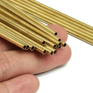 May include: A collection of small, hollow brass tubes. The tubes are cylindrical and have a shiny, golden appearance. Some are held in a hand, while others are in the background. These tubes are likely used for crafts or jewellery making.