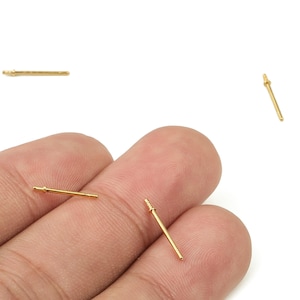 May include: Close-up of several small, gold-colored earring posts. The posts are straight with a small, textured end and a flat, round end. The posts are on a white background, with some held in a person's fingers.