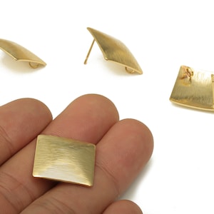 May include: Gold-coloured metal earrings. The earrings have a square shape with a textured surface. Two earrings are post-style, and one is held in a hand. The earrings have a minimalist, geometric design.