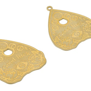 May include: Two gold-colored metal charms shaped like a witch's pyramid or a Ouija board planchette. Each charm has a circle in the center, a floral design, and the words "Good Luck" inscribed on the surface.