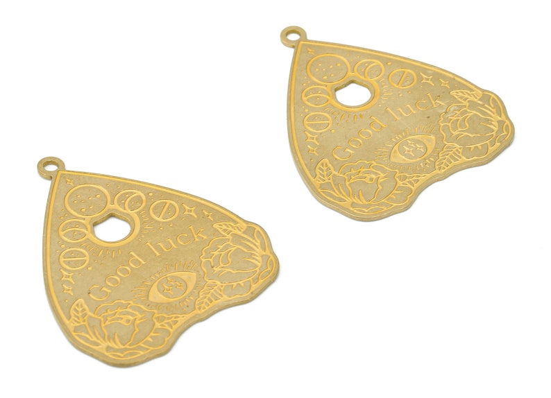 May include: Two gold-colored metal charms shaped like a witch's pyramid or a Ouija board planchette. Each charm has a circle in the center, a floral design, and the words "Good Luck" inscribed on the surface.