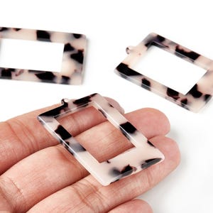 May include: Three rectangular resin charms with a tortoiseshell pattern of black and cream. Each charm has a rectangular opening in the center and a small loop at the top for attaching to jewelry. The charms are shown on a white background.