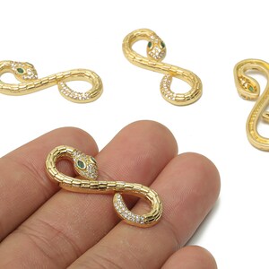 Puede incluir: Encantos dorados en forma de serpiente con detalles de cristal verde y transparente. Los amuletos tienen un diseño serpentino, uno de ellos se sostiene en una mano. Los ojos de la serpiente son verdes y el cuerpo está adornado con cristales transparentes.