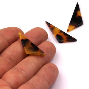 May include: Three triangular, tortoiseshell-patterned pendants. One is held in a hand, while two others are placed on a white surface. The pendants have a hole at the top, and are a mix of black, brown, and amber.
