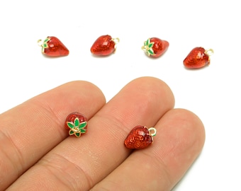 Puede incluir: Pequeños dijes de fresas rojas con lazos dorados para hacer joyas. Las fresas tienen hojas verdes y un acabado brillante y texturizado. Varios dijes están esparcidos sobre un fondo blanco, algunos de ellos sostenidos en una mano.