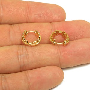 Op de afbeelding: Een paar goudkleurige oorringen. Elke oorring heeft een gedetailleerd bladerontwerp rond de ring. De oorbellen worden op een hand getoond tegen een witte achtergrond.