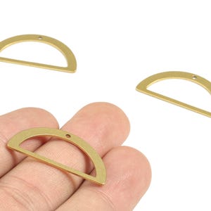 May include: Three brass-coloured, half-circle shaped charms. Each charm has a small hole at the top for stringing. One charm is held in a hand. These charms are suitable for jewellery making or other craft projects.
