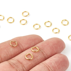 May include: A collection of small, gold-colored jump rings, some held by a hand. The rings are circular and appear to be made of metal. They are scattered on a white surface, suggesting their use in jewelry making or crafts.