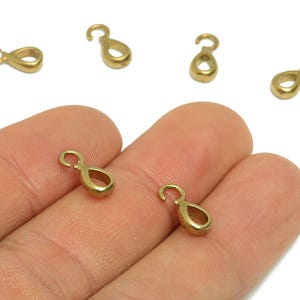 以下が含まれることがあります： 小さな金色のジュエリークラスプのクローズアップ。 クラスプは、上部に小さなループが付いた無限大の記号の形をしています。 いくつかのクラスプは人の手にあります。