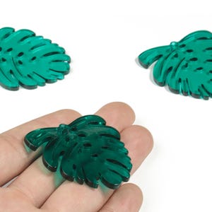 Puede incluir: Tres colgantes transparentes en forma de hoja de Monstera, de color verde esmeralda. Uno se sostiene en una mano, mientras que los otros dos se muestran por separado. Las hojas tienen un acabado brillante y están diseñadas con venas y agujeros detallados.
