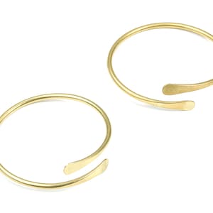 Può includere: Due braccialetti aperti dorati con un design semplice e minimalista. Ogni braccialetto ha un unico pezzo piatto e curvo che si estende dal cerchio principale.