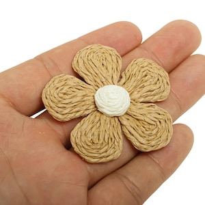 Peut inclure: Une fleur tissée à cinq pétales et un centre blanc. Les pétales sont de couleur beige clair et le centre est blanc. La fleur est faite d'un matériau tissé et est tenue dans une main.