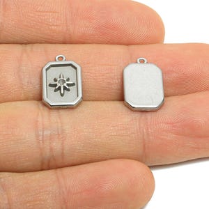 Puede incluir: Dos colgantes rectangulares plateados. Uno presenta un diseño de estrella dentro de un marco geométrico. El otro colgante es un rectángulo liso. Ambos tienen un pequeño lazo en la parte superior para sujetarlos a una cadena u otra joyería.
