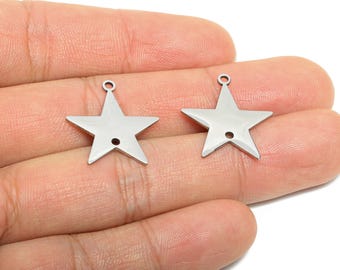 Puede incluir: Dos colgantes plateados en forma de estrella. Cada colgante tiene un pequeño agujero en la parte inferior y un lazo en la parte superior para sujetar. Los colgantes son lisos y brillantes, reflejando la luz.