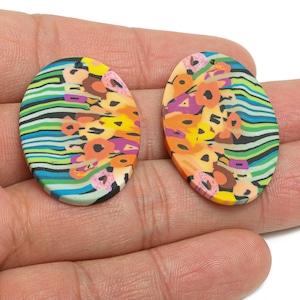 May include: A pair of oval polymer clay earrings. Each earring displays a vibrant abstract design with orange, yellow, pink, and black floral patterns, set against blue, green, and black striped backgrounds.