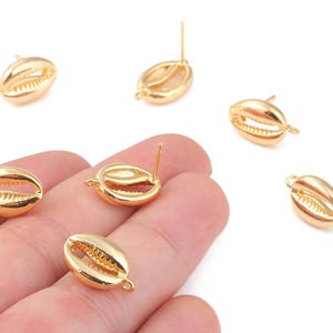 Könnte beinhalten: Mehrere goldfarbene Ohrringkomponenten in Form von Kaurischalen. Die Ohrringe haben einen Stift zum Einsetzen und eine Schlaufe zum Anbringen zusätzlicher Elemente. Die Muscheln sind mit inneren Rippen und einer glatten, polierten Außenseite versehen.