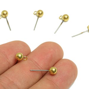 May include: Gold-colored ball stud earrings with a silver post and a small loop. Several earrings are scattered on a white background, with two held in a hand. The earrings have a simple, minimalist design.