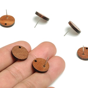 Op de afbeelding: Een verzameling houten oorstekers. De ronde oorbellen hebben een donkerbruine afwerking en een zilveren pin. Eén oorbel heeft een klein gaatje aan de bovenkant. Verschillende oorbellen liggen op een wit oppervlak.
