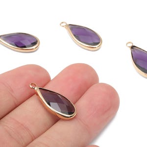 May include: Four teardrop-shaped pendants with a faceted purple gemstone set in a gold-colored metal frame. One pendant is held in a hand. The pendants have a small loop at the top for attaching to a chain or other jewelry.