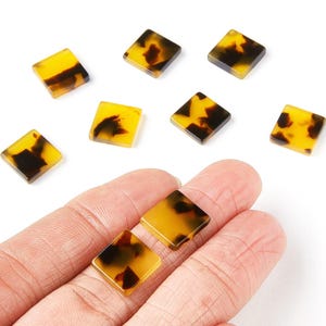 May include: Square-shaped beads with a tortoiseshell pattern. The beads are a mix of yellow and dark brown, with a glossy finish. Several beads are scattered on a white surface, and two are held in a hand.
